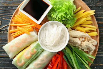Wooden board with spring rolls and ingredients on wooden background