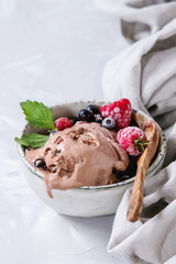 Melting homemade chocolate ice cream with frozen berries blueberry, raspberry, mint served in white bowl with olive wood spoon and textile linen over gray concrete background. Close up