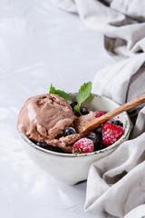 Melting homemade chocolate ice cream with frozen berries blueberry, raspberry, mint served in white bowl with olive wood spoon and textile linen over gray concrete background. Close up