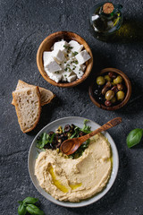 Obraz na płótnie Canvas Homemade traditional spread hummus with chopping olives and herbs on blue plate, served with bread, olives, feta cheese, olive oil, spoon on black texture background. Mediterranean snack. Flat lay