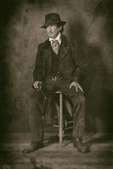 Fototapeta na wymiar Classic wet plate photo of vintage 1900 western mature man with revolver sitting on wooden stool.