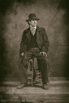 Classic wet plate photo of vintage 1900 western mature man with revolver sitting on wooden stool.
