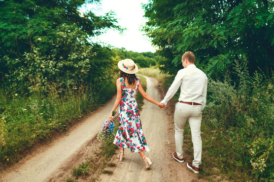 Happy young couple walking with arm around on trail in forest. Lovers holding hands. Girl in a straw hat carries a bouquet