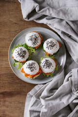 Mini rice sushi burgers with smoked salmon, green salad and sauces, black sesame served on gray plate with textile linen over wood background. Modern healthy food. Top view with space