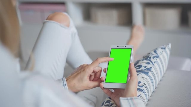 Young Woman in white jeans laying on couch uses smartphone with pre-keyed green screen. Few types of gestures - scrolling up and down, tapping, zoom in and out. Perfect for screen compositing
