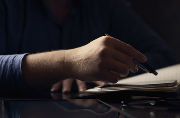 Close-up of a man sitting in a dark-colored workplace and working the pen on the phone and a diary