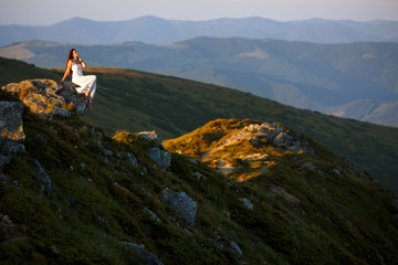 Beautiful woman in a long white dress in the mountains. Young woman sitting on a rock. Hair blowing in the wind