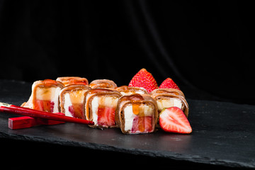 new trend in the Japanese cuisiune. Sweet sushi rolls in pancake with cream cheese, strawberry and banana throwing with jam, dark background