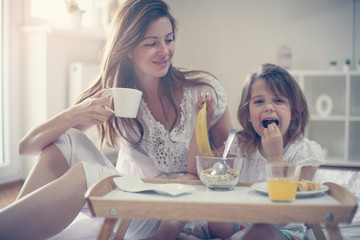 Mother with her little daughter having breakfast in the bed. Mother and her daughter enjoying a healthy breakfast together. Little girl eating banana.