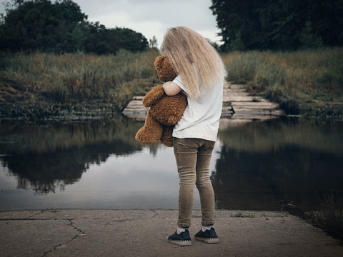 Sad Child Hugging A Toy Teddy Bear. Field, River Bank