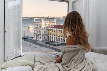 Woman sitting on the bed in the hotel room and looking at view from window at Lisbon city streets...