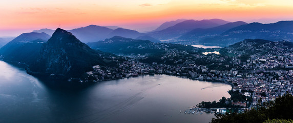 Fototapeta na wymiar Aerial view of the lake Lugano surrounded by mountains and evening city Lugano on during dramatic sunset, Switzerland, Alps. Travel