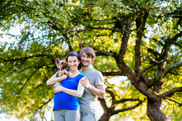 Pregnant sporty couple holding yorkshire terrier, smilling and looking at camera over beautiful green tree background. Parenthood, sport and healthy life concept.
