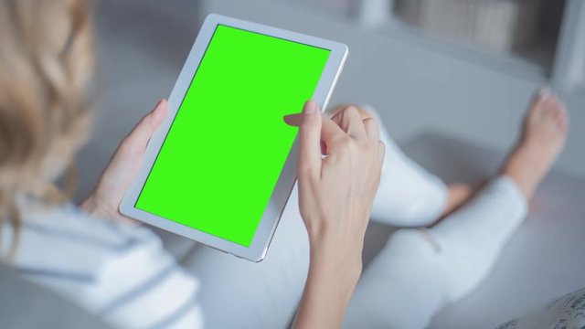 Young Woman in white jeans sitting on couch uses Tablet with pre-keyed green screen. Few types of gestures - scrolling up and down, tapping, zoom in and out. Perfect for screen compositing