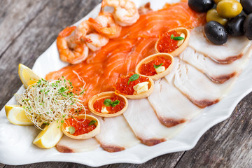 Seafood platter with salmon slice, pangasius fish, red caviar, shrimp, decorated with olives and lemon on wooden background close up. Mediterranean appetizers. Top view