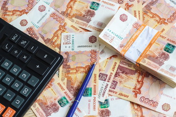 Concept planning expenses: calculator and ball-point pen and a pack of half a million Russian rubles in the banking package lying on the background of scattered banknotes