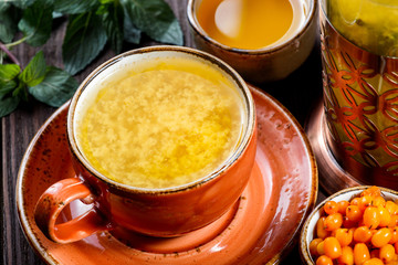 Tea with sea Buckthorn in cup with saucer, served with vintage teapot, fresh berries, honey, mint  on dark wooden background