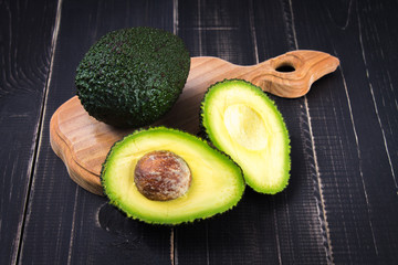 Cut and whole avocado on a wooden board on a black wooden background.