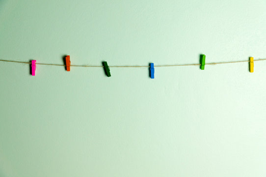 Small multi color wooden clips hanging with brown rope on light green wall.