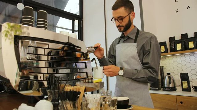 Handsome barista in the glasses preparing cup of coffee for customer in modern coffee shop. View from the side.