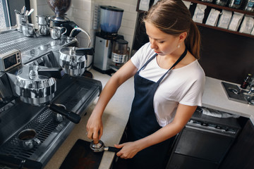 Young Caucasian woman barista using tamper to press ground coffee into portafilter to make espresso hot drink. Small local business, work in cafe.