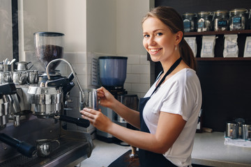 Portrait of  happy beautiful young Caucasian smiling woman barista holding milk jug for making coffee using coffee machine. Small local business with organic hot drinks products. Toned with filters.