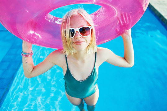pink floaty with blonde teen girl in white sunglasses wearing a one piece in water wet swimming pool