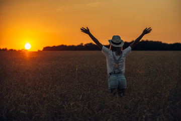 Young girl with outspread hands standing standing in the wheat field at sunset