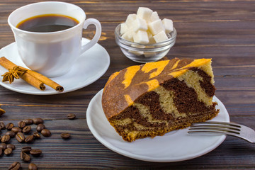 Delicious cake on a plate with a fork. White cup with black coffee and cinnamon sticks near it. Coffee beans and a bowel with sugar cubes on dark wooden table