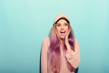  beautiful young woman in pink shirt posing with hand on cap and looking away. Three quarter length studio shot on turquoise background