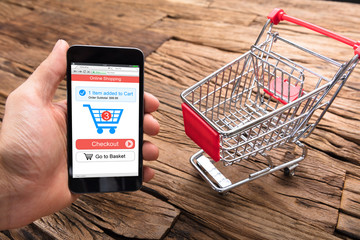 Businessman's Hand Holding Smartphone By Shopping Cart On Table
