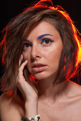 Unretouched portrait of a young pretty caucasian brunette girl posing in a studio on black background with bright orange backlight. She has messy hair and bright blue eye liner and beige lip gloss. - 167408999