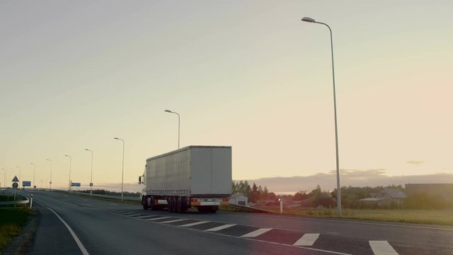 Semi-Truck with Cargo Trailer Drives on a Highway. Static Camera Follows Him. In the Background Rural Area with Rising Sun. Shot on RED EPIC-W 8K Helium Cinema Camera.