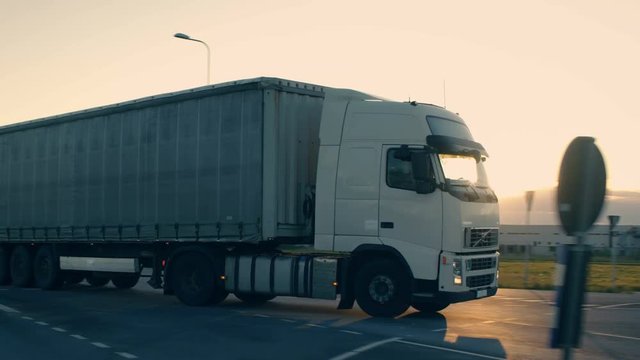 Follow-up Shot of a Semi-Truck with Cargo Trailer Moving on a Highway. White Truck Drives Through Industrial Warehouse Area in Early Hours of the Morning. Shot on RED EPIC-W 8K Helium Cinema Camera.