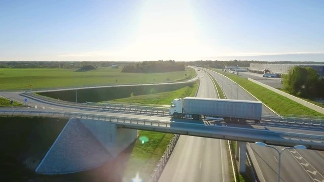 Aerial View of White Semi Truck with Cargo Trailer Passing Highway Overpass/ Bridge. Eighteen Wheeler is New, Loading Warehouses are Seen in the Background. Shot on RED EPIC-W 8K Helium Cinema Camera.