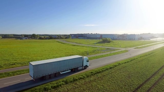 Aerial View of White Semi Truck with Cargo Trailer Moving on the Highway. In the Background Warehouses and Rural Area, Sun Shines. Shot on RED EPIC-W 8K Helium Cinema Camera.