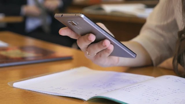 Girl holding a steel color mobile phone sits at a school desk typing the text using this gadget. Close-up