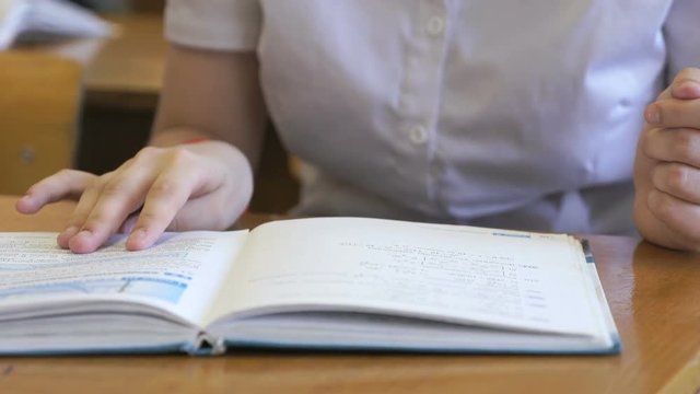 Student dressed in white shirt sitting at school desk flips the pages of the school book. Close up