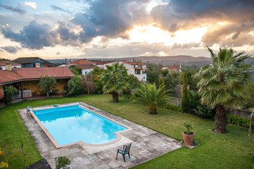 View on a luxury villa and a bright blue pool on a lawn. Shot during a sunset with a dramatic cloudy sky and golden rays shining through the clouds. Shot in Portugal, Villa Nova de Poiares.