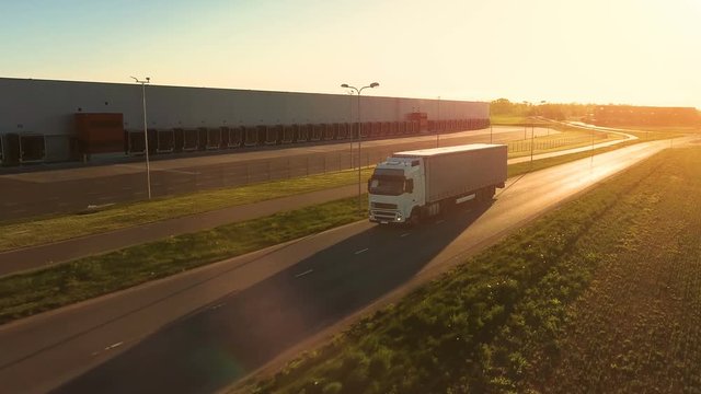 Aerial View of White Semi Truck with  Cargo Trailer Moving on the Highway. In the Background Warehouses and Rural Area, Sun is Setting. Shot on RED EPIC-W 8K Helium Cinema Camera.