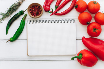 Notebook with a recipe. Red pepper, rosemary, tomatoes and green chilies on a white wooden background.