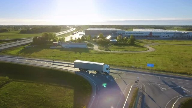 Aerial View of White Semi Truck with Cargo Trailer Passing Highway Overpass/ Bridge. Eighteen Wheeler is New, Loading Warehouses are Seen in the Background. Shot on RED EPIC-W 8K Helium Cinema Camera.