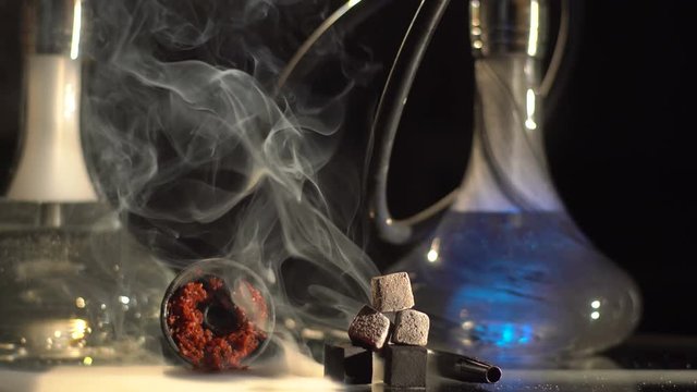 Horizontal view of the composition consisting of the embers and hookahs on the table is surrounded by thick smoke. The modern hookah is changing the colour.