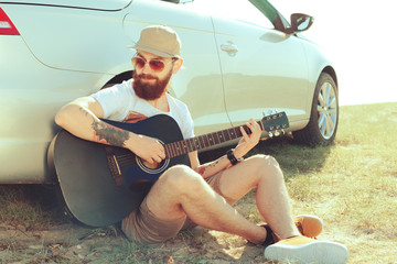 Bearded man playing guitar outdoors near car