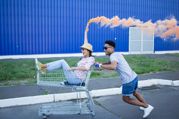 Young beautiful couple in love, boyfriend and girlfriend ride on shopping cart with orange smoke...