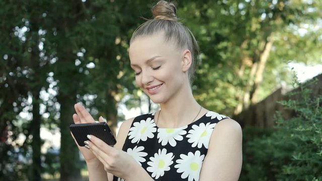 Pretty girl in floral shirt standing in the park and browsing internet on smartphone, steadycam shot
