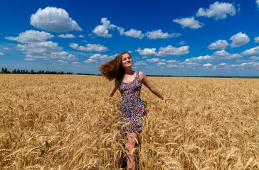 Naklejka premium Beautiful young woman walking in golden wheat field with cloudy blue sky background, free space. Liberty, peace of mind concept. Girl in spikes of ripe wheat field under blue sky