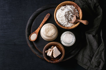 Fototapeta na wymiar Rye and wheat sourdough in glass jars, fresh and instant yeast, olive wood bowl of flour for baking homemade bread. With spoon, serving board, textile over black burned wooden background. Top view
