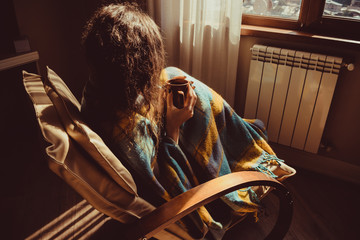 Winter concept. Young woman sitting in comfortable modern chair near radiator with mug of tea wrapped in warm plaid blanket. Natural light. Warm atmosphere. Cup Hot Beverage. Heating season. Cosy home