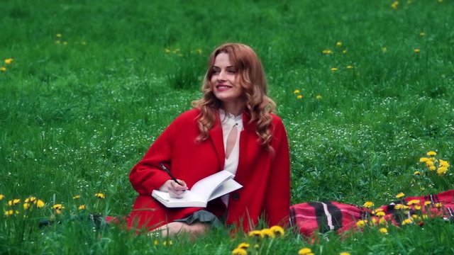 Happy beautiful adult woman sits on the grass, writes in notebok, smiles. Outdoor, full body view